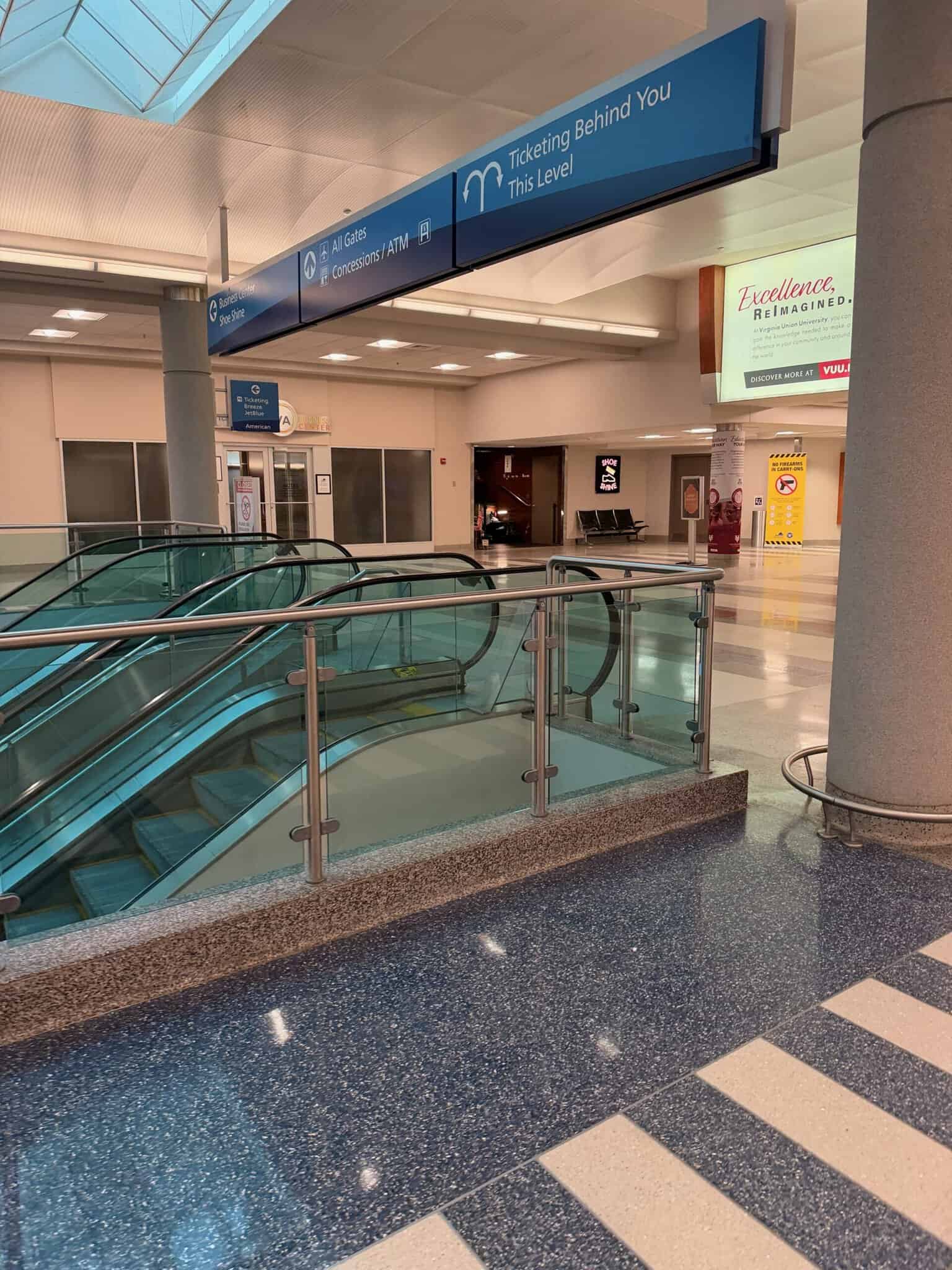 Empty escalators at RIC airport under blue overhead signs pointing to ticketing, all gates, concessions and ATM