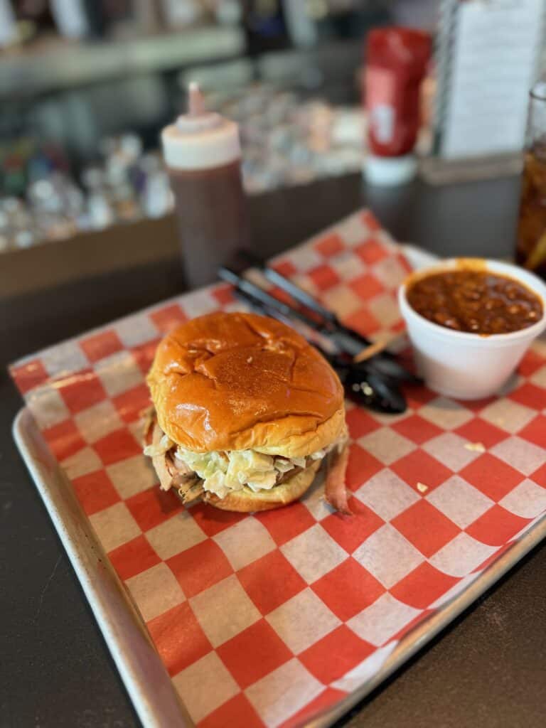 A pulled pork BBQ sandwich with coleslaw and a side of baked beans on a red-checkered tray at a DFW airport restaurant - aviation photography by Anthony Yandow