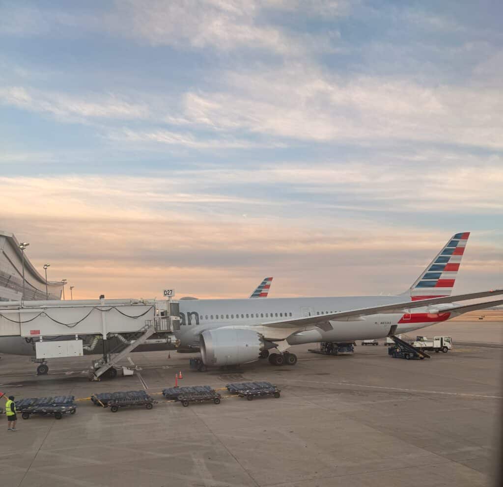 An American Airlines widebody jet at Gate D27 in DFW Terminal D at dusk, with the modern curved terminal building visible - aviation photography by Anthony Yandow