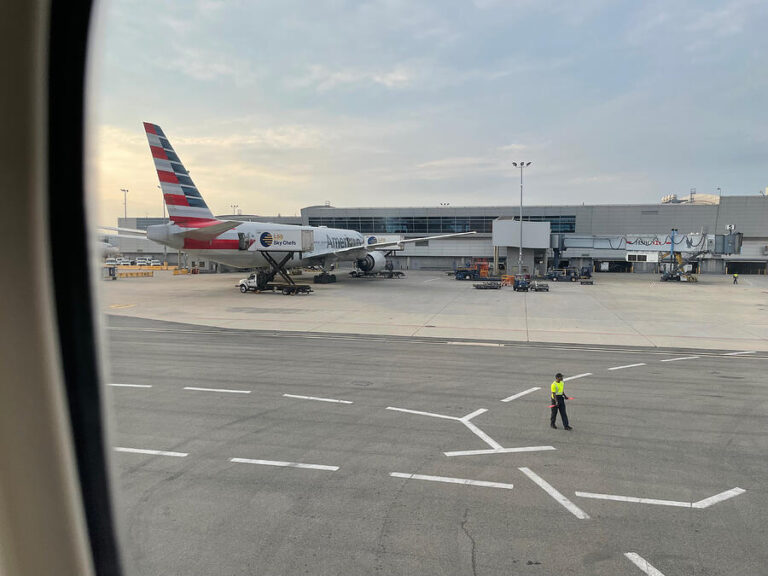 A Boeing 777 parked at a JFK terminal gate with a ground crew member walking nearby