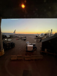 A Boeing 787 and 777 parked at DFW at sunrise with ground crew vehicles and cargo on the ramp